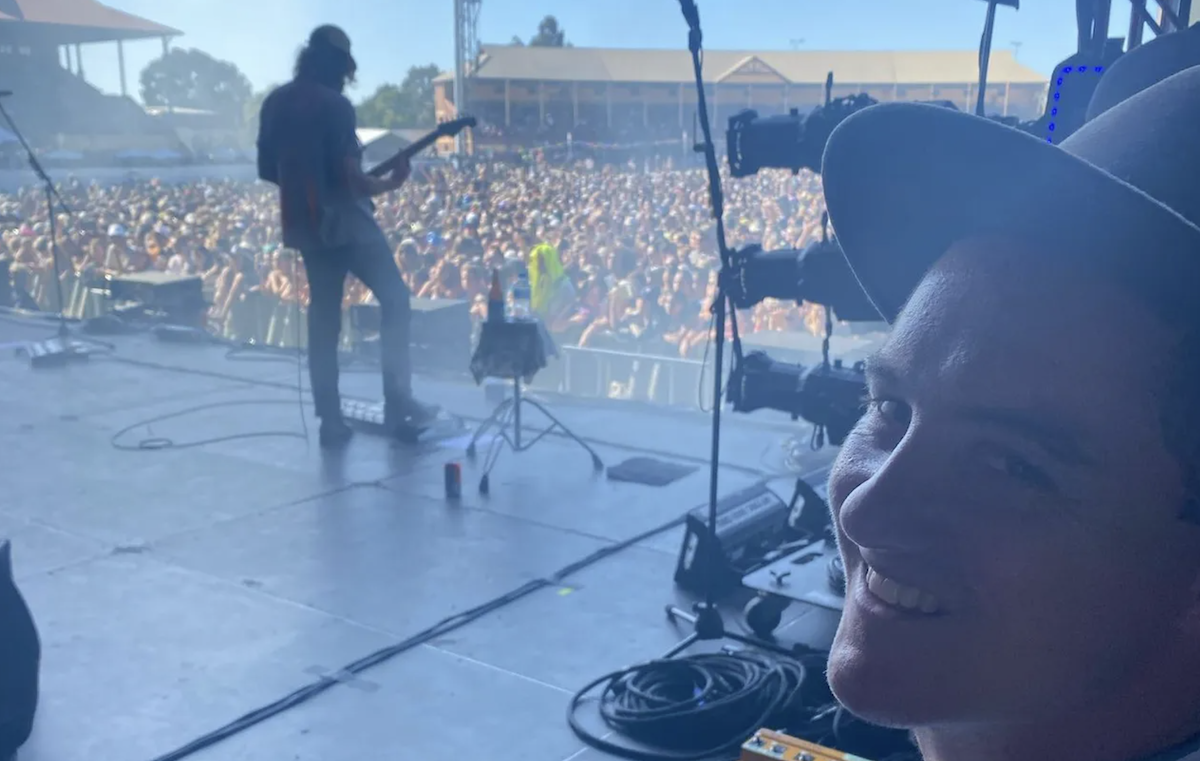 ABC reporter Charles Brice smiling backstage at a music festival, with a guitarist performing on stage in front of a large outdoor crowd behind him.